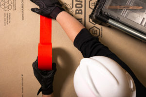 Bovenaanzicht van een man met een harde hoed en handschoenen die rode Pro tape-ducttape aanbrengt op twee vellen ramboard voor zware vloerbescherming