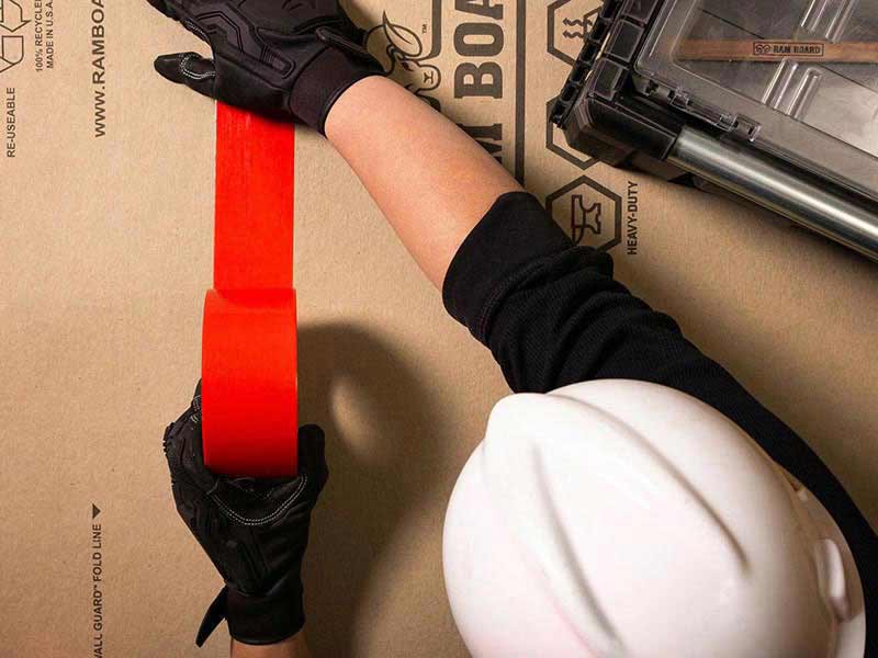 Top down view of a man in a hard hat and gloves applying red Pro tape duct tape to two sheets of ram board heavy duty floor protection